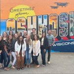 A photo of the E-Power Marketing team in front of the Oshkosh Greetings Tour Mural.
